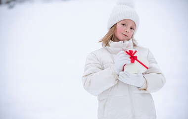 A joyful little girl in white winter clothes stands in front of the snow and holds a heart-shaped box