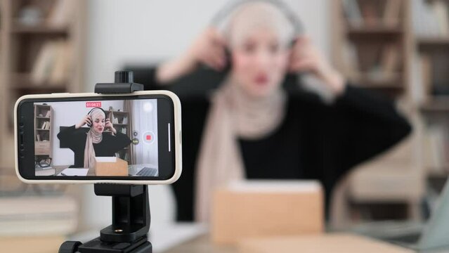 Pleasant arab woman filming video on modern phone camera while opening parcel box with new wireless headphones. Concept of people, technology and blogging.
