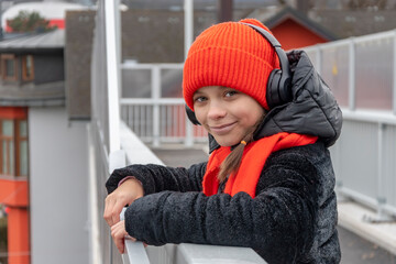 Street portrait of a happy 11-year-old girl in a red hat and headphones on a pedestrian bridge.