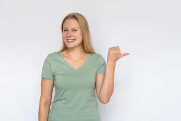 Cheerful young woman pointing thumb aside. Portrait of happy Caucasian female model with fair hair in green T-shirt looking at camera, smiling and showing ads. Advertising concept
