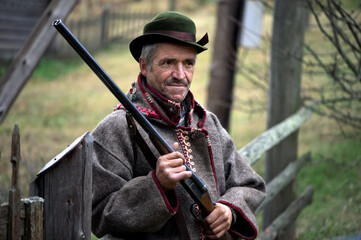  A hunter with a gun in his hands in hunting clothes stands near the forest hut.