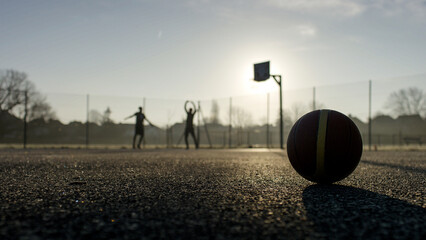 Wide angle shot of basketball in the foreground as two silhouette people workout in the background on an outdoor basketball court