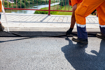 Fototapeta premium Workers apply black bitumen membrane strips for waterproofing roads and bridges to the curb of the sidewalk
