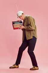 Young emotive man in casual clothes posing with vintage radio set, music player over pink studio background. Listening to news. Concept of emotions, facial expression, lifestyle, retro fashion. Ad
