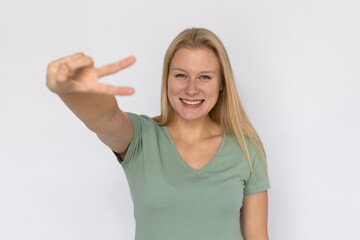 Fototapeta premium Excited young woman showing victory gesture. Portrait of happy Caucasian female model with fair hair in green T-shirt looking at camera, smiling and greeting someone. Success, optimism concept
