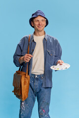 Naklejka premium Positive mood, inspired look. Portrait of young man in jeans clothes, painter, artist posing over blue studio background. Concept of emotions, facial expression, lifestyle, hobby, occupation. Ad