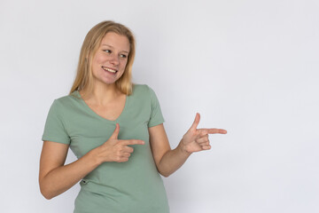 Happy young woman pointing fingers aside. Portrait of pleased Caucasian female model with fair hair in green T-shirt looking away, smiling, showing ads or greeting someone. Advertising concept