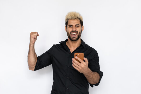 Joyful businessman using smartphone. Male Caucasian model with brown eyes, ombre painted hair and beard in black shirt raising his fist with face of winner. Success concept