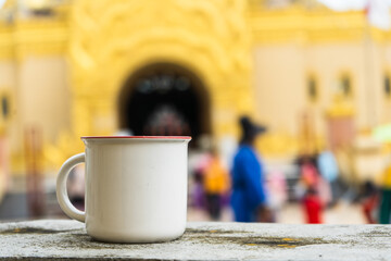 An enamel mug standing on the top of a fence with out of focus golden temple and some people