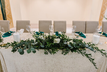 Wedding set up, dinner table reception. Flower composition with eucalyptus leaves and candles in the center of the table. A plate with a green cloth towel, knives and forks next to the plate.  Closeup