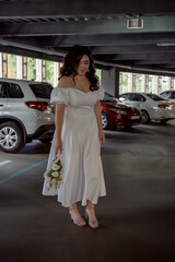 Bride in white airy dress with bouquet of roses in hands stands in parking lot, behind a row of expensive luxury cars. The Bride in Urban Style