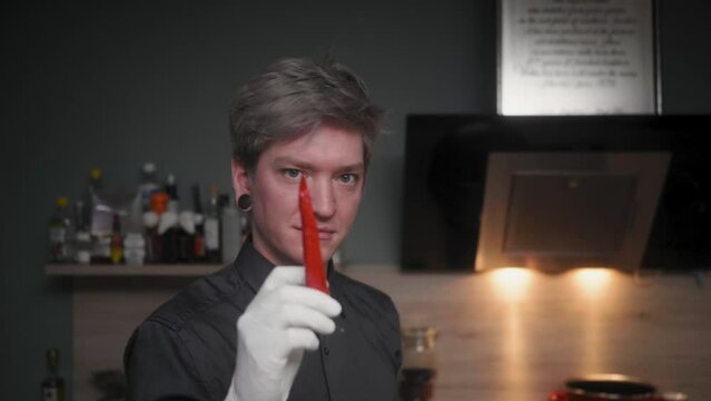 Professional attractive male chef in an elegant black shirt with an alternative look (tattoos, colored hair) biting off a red chili while winking into camera 
A modern, fancy looking kitchen, Slow Mo