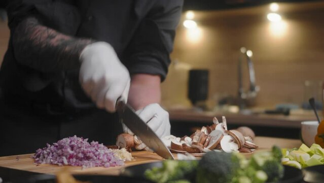 Mushrooms being chopped on a wooden board by a young male chef in an elegant black shirt with tattoos wearing white gloves. Fresh vegetables around in a modern, kitchen, Slow Motion cinematic sliding