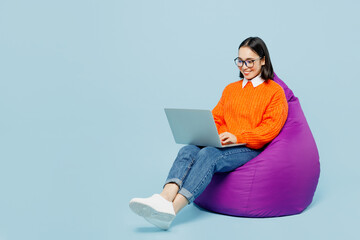 Full body young IT woman of Asian ethnicity wear orange sweater glasses sit in bag chair work hold...