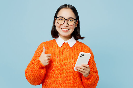 Young Smiling Happy Fun Woman Of Asian Ethnicity Wear Orange Sweater Glasses Hold In Hand Use Mobile Cell Phone In White Case Show Thumb Up Isolated On Plain Pastel Light Blue Cyan Background Studio.