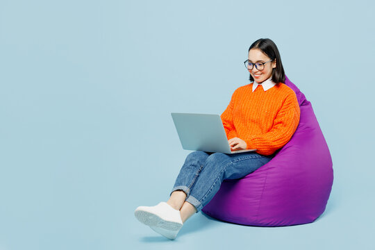 Full Body Young IT Woman Of Asian Ethnicity Wear Orange Sweater Glasses Sit In Bag Chair Work Hold Use Laptop Pc Computer Isolated On Plain Pastel Light Blue Cyan Background. People Lifestyle Concept.