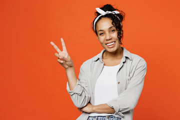 Young cheerful fun smiling happy woman of African American ethnicity she wears grey shirt headband showing victory sign isolated on plain orange background studio portrait. People lifestyle concept.