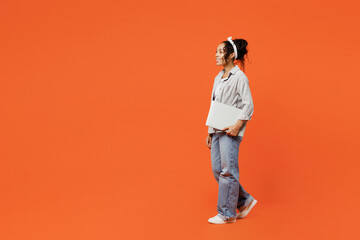 Full body side profile view student young woman of African American ethnicity she wears grey shirt headband hold closed laptop pc computer walk go isolated on plain orange background studio portrait.