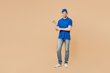 Full body smiling delivery guy employee man wear blue cap t-shirt uniform workwear work as dealer courier point index finger aside on area isolated on plain light beige background. Service concept. - obrazy, fototapety, plakaty