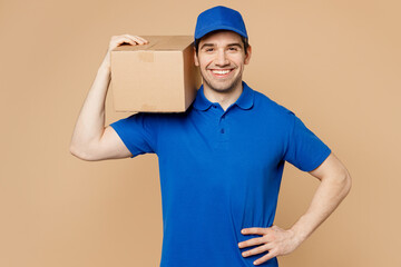 Professional smiling delivery guy employee man wear blue cap t-shirt uniform workwear work as dealer courier hold cardboard box on shoulder isolated on plain light beige background. Service concept.