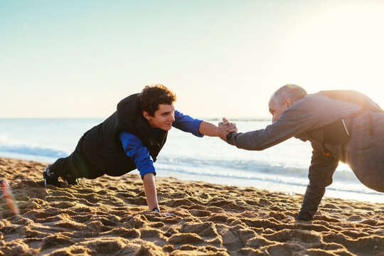 Side view of father and son doing push-ups while holding hands against clear sky at beach during sunset