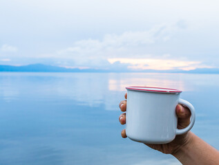 A hand holding an enamel mug while showing the out of focus scenery at the background