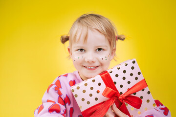 Cheerful little girl in pink sweatshirt with red hearts pattern holding gift box with red ribbon isolated over yellow background.