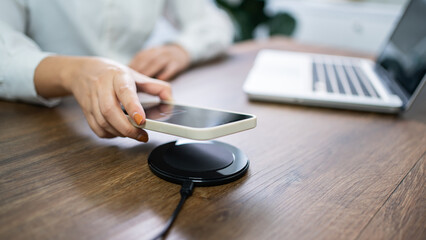 Charging mobile phone battery with wireless charging device in the table. Smartphone charging on a charging pad. Mobile phone near wireless charger Modern lifestyle technology concept. - obrazy, fototapety, plakaty