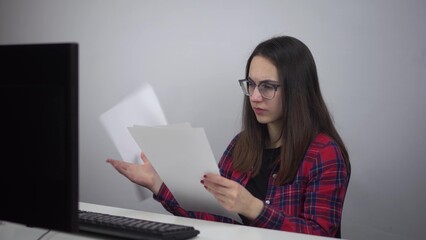 Naklejka premium A young IT lady reads documents and throws them out of anger. A woman is sitting at the workplace in the office. Girl in a red shirt and glasses.
