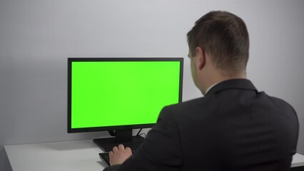 A young businessman is sitting in front of a computer with a green screen. A man in a suit works for a PC. Screen with chroma key.