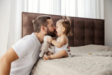 A father is rubbing noses with his little girl while she is sitting on a bed and speaking something. - obrazy, fototapety, plakaty