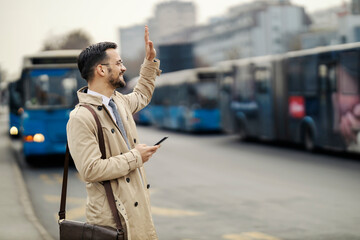 A happy young man in smart casual is using phone and calling a cab on a bus station.