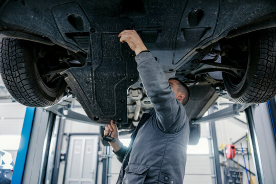 A car workshop worker is repairing car while standing under it at mechanic's shop.