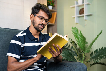 serious young indian student reading book during free time at home - concept of education, hobbies...