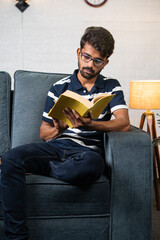 Vertical shot of serious young indian student reading book during free time at home - concept of education, hobbies and hard worker.