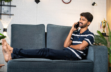 Obraz premium wide shot of Happy smiling young man talking on mobile phone on sofa at home - concept of communication, technology and relaxed conversation.
