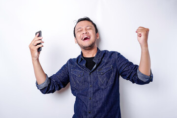 A young Asian man with a happy successful expression wearing blue shirt and holding his phone,...