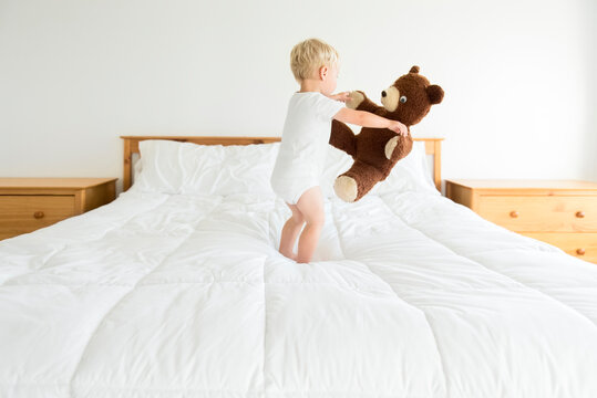 Cute boy playing with teddy bear while standing on bed