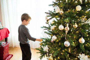 Young boy stands placing ornament on Christmas tree during holiday