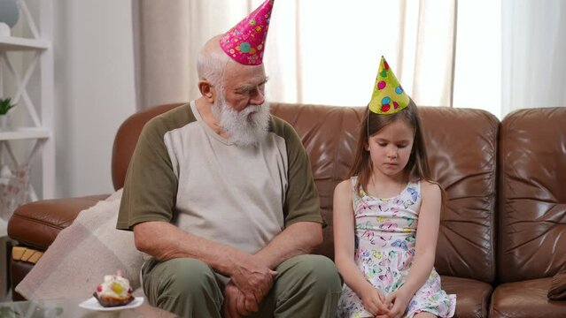 Sad old man sitting with bored charming girl in party hat on couch looking at birthday cupcake. Portrait of upset Caucasian granddaughter on holiday with grandfather at home. Slow motion