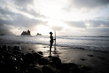 Silhouette of a surfer holding surfboard and looking at the sky at the beach.