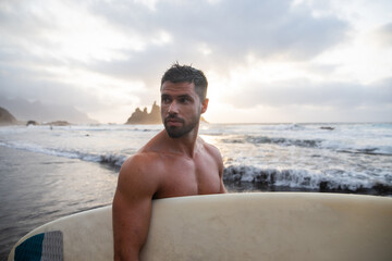Portrait of a surfer holding his surfboard and looking to the side at the beach.