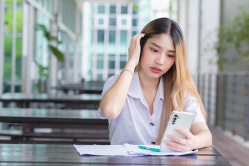 Portrait of cute Asian Thai girl student in uniform is sitting work smiling happily and confidently while using smartphone in the building at university with background.