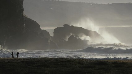 Power of nature. Two people watch huge waves crashing against the reef from the beach.