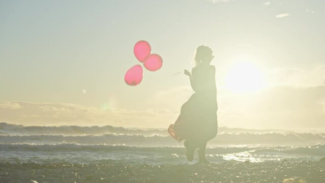 Young woman walks along beach holding three red balloons as sun sets behind her