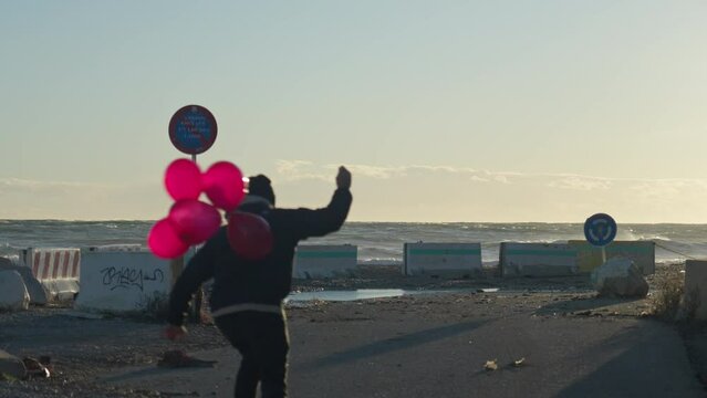 Skateboarder rides towards the sea holding bunch of red balloons as some escape in the wind