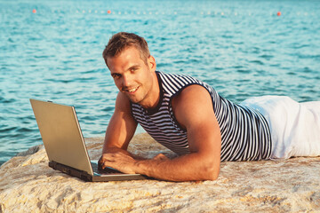 Man with the computer on the rocky beach wearing a sleeveless sailor shirt 