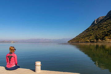 Woman sitting on pier with panoramic view of Lake Skadar National Park in autumn seen from Godinje, Bar, Montenegro, Balkans, Europe. Stunning travel destination in Dinaric Alps near Albanian border