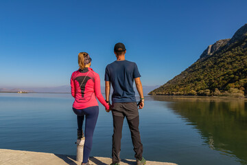 Fototapeta premium Couple standing holding hands on pier with panoramic view of Lake Skadar National Park in autumn seen from Godinje, Bar, Montenegro, Balkans, Europe. Stunning travel destination in Dinaric Alps