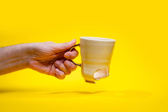 Man holding broken tea cup isolated on yellow background. Close up of arm holding cracked coffee mug